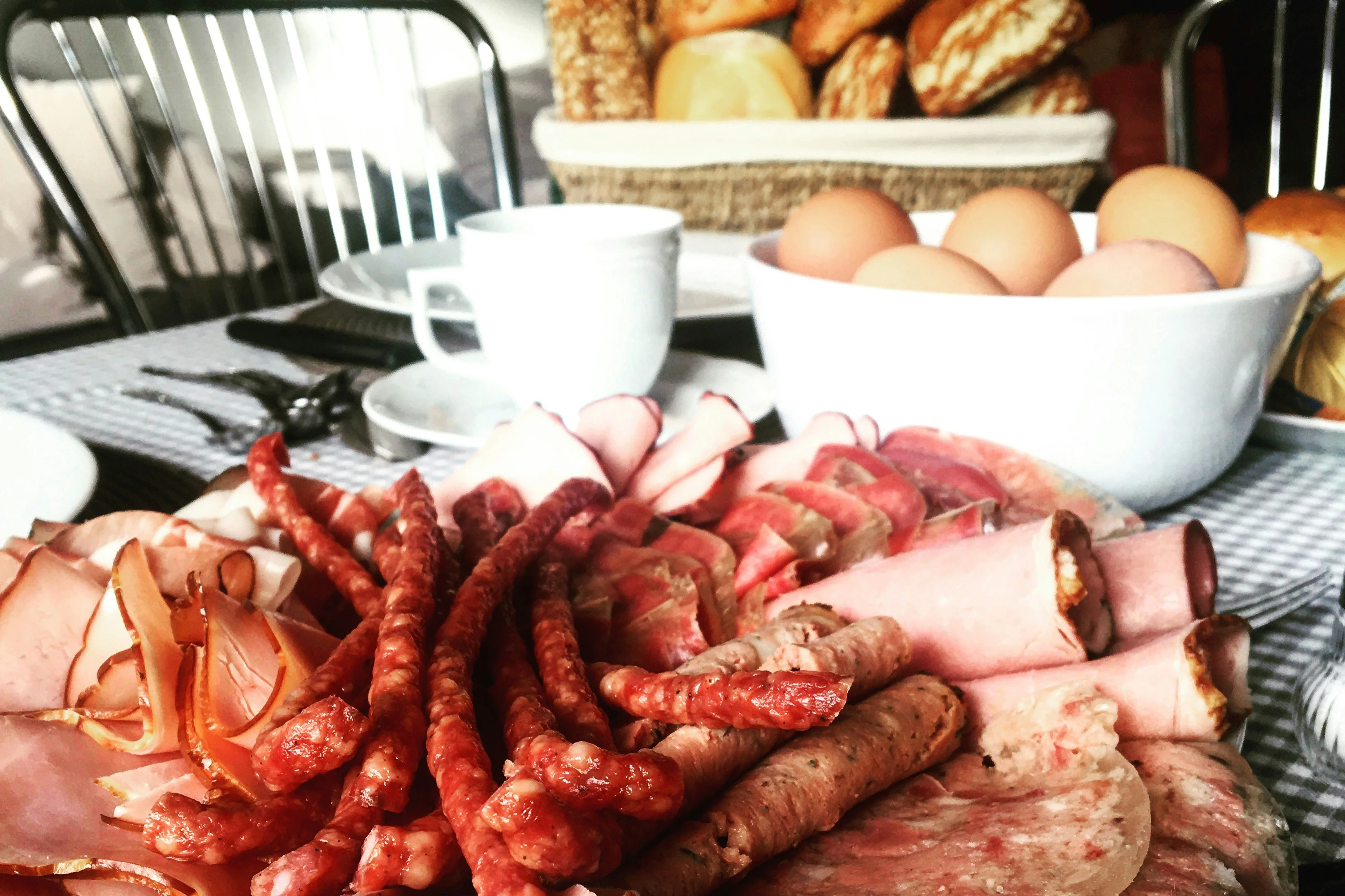 A German breakfast spread: a platter of sliced and whole sausages, a bowl of boiled eggs, a cup of coffee, and a basket of fresh bread rolls.